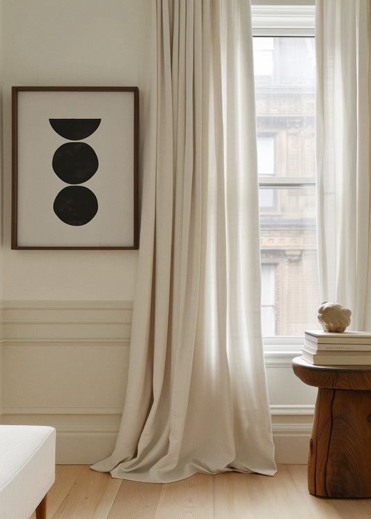 Minimalist wall art with black geometric shapes titled "Boulders" in a wooden frame, styled in a soft and neutral living room with flowing curtains and natural light.