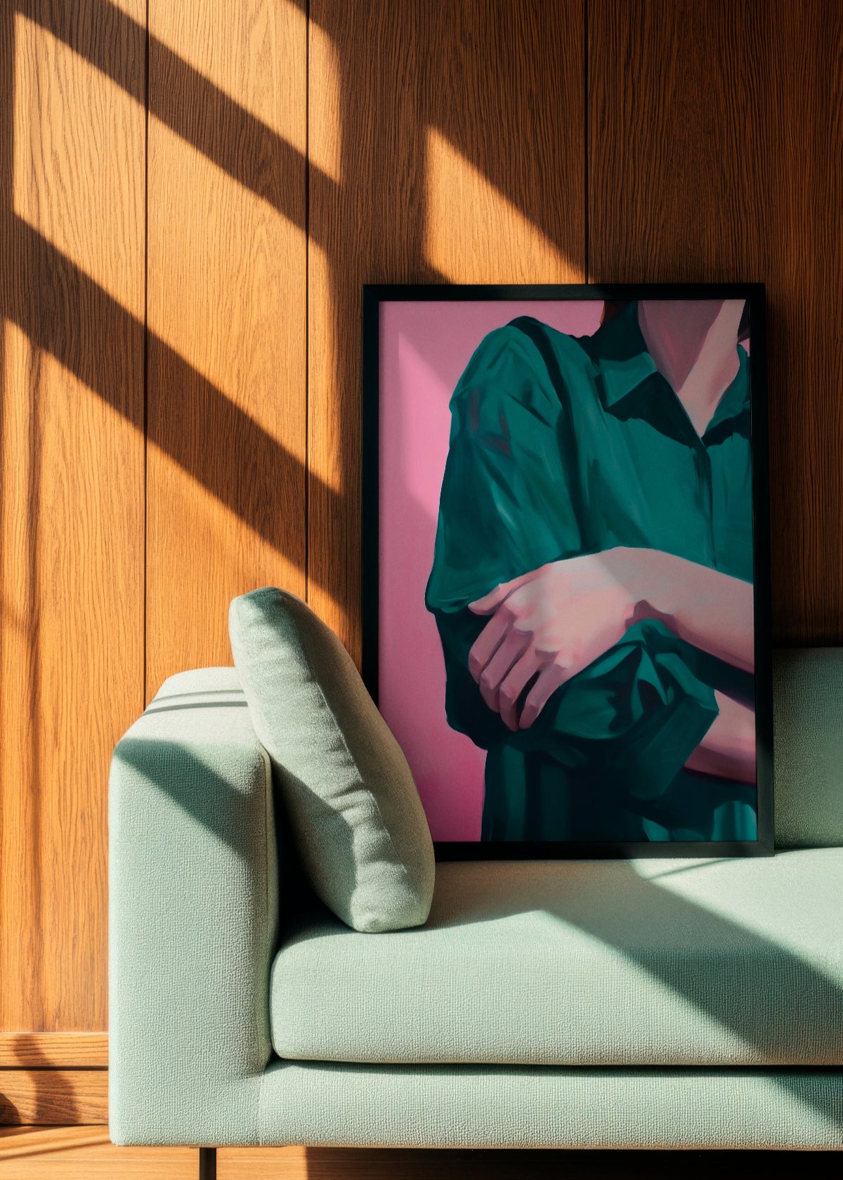 Silent Pose poster featuring a serene depiction of folded arms in a green shirt against a soft pink background, framed in black and leaning against a wooden-paneled wall beside a light green sofa. The artwork conveys calmness and introspection, perfect for minimalist and modern interiors.