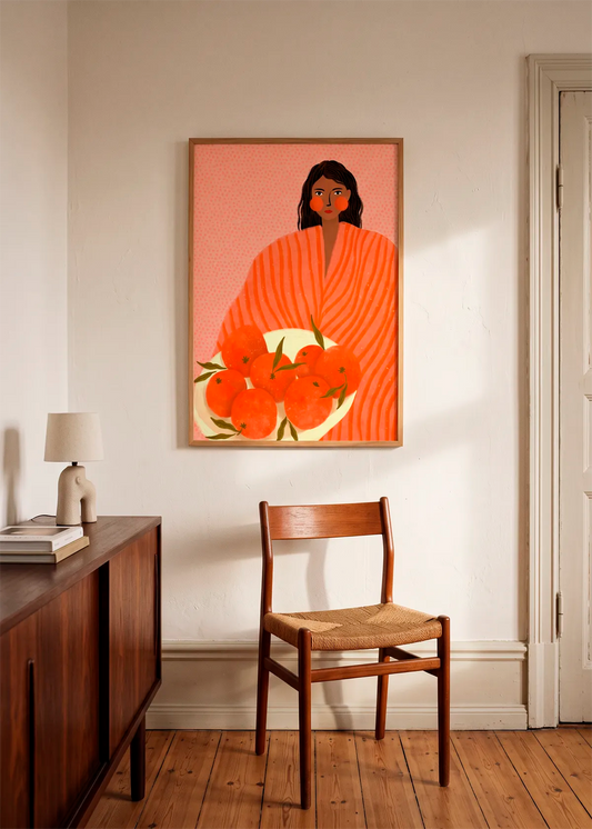 Woman Holding Oranges by Bea Müller – framed art poster featuring a woman in orange holding a bowl of oranges, styled in a warm neutral interior with wooden furniture.
