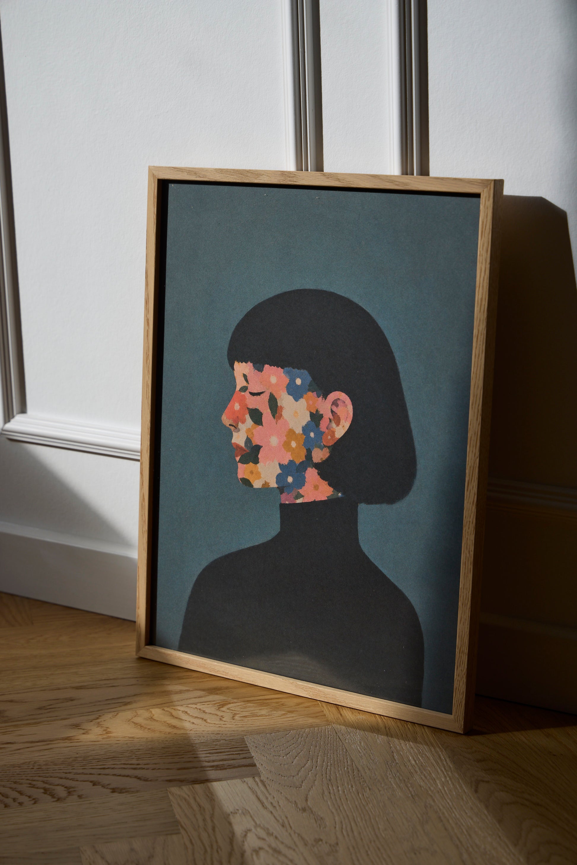 A Face I Once Knew poster featuring a woman’s profile with colorful flowers in muted pink, blue, orange and green tones, styled in a modern Scandinavian interior with an oak frame.