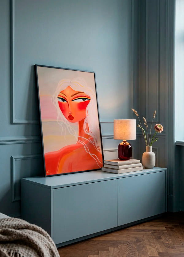 Blonde Sunset Gaze by Amy Straley – framed portrait art poster featuring a woman with blonde hair, warm coral skin tones and a soft sunset palette, styled on a blue sideboard in a moody blue interior.