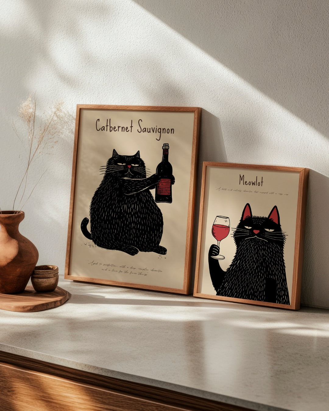 Two playful cat posters titled "Catbernet Sauvignon" and "Meowlot" displayed together in oak frames, adding humor and charm to a kitchen or dining space.