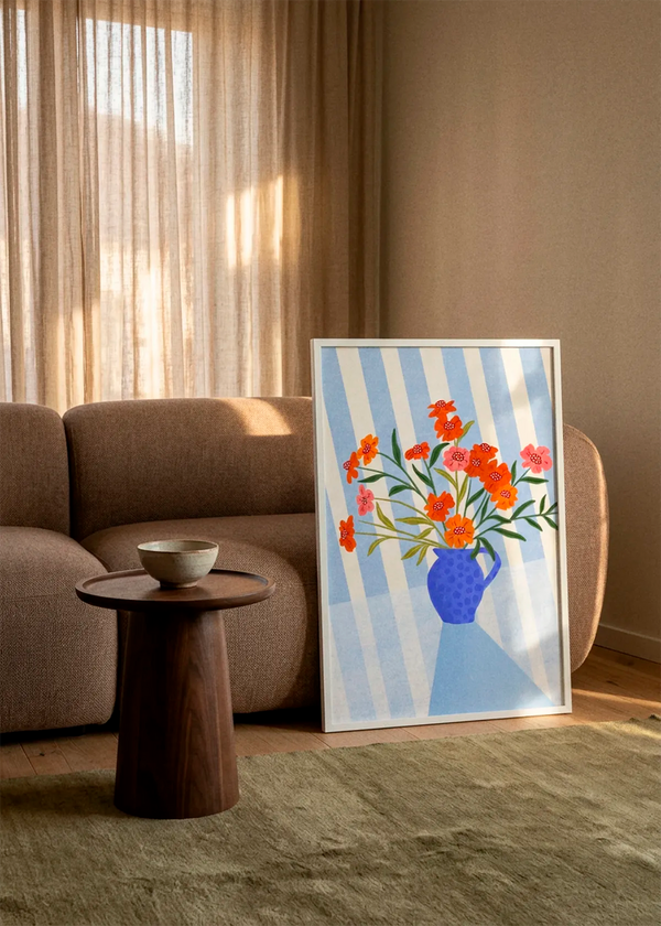 Cheerful Floral Pitcher by Bea Müller art print featuring orange and pink flowers in a vivid blue pitcher against soft blue and cream stripes, displayed in a white frame in a warm Scandinavian living room.