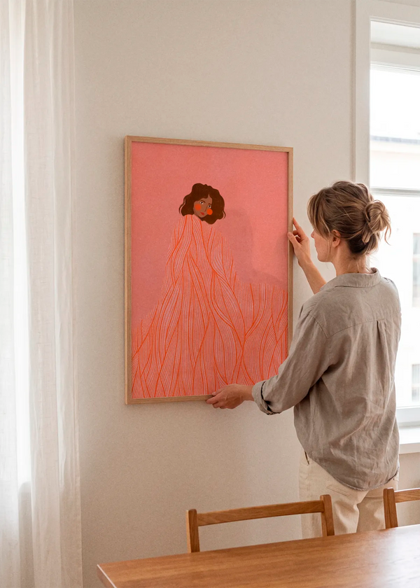 Coral Woman Dreaming by Bea Müller art print featuring a woman wrapped in a flowing coral striped garment against a soft pink background, displayed in a light wood frame in a bright Scandinavian dining room.