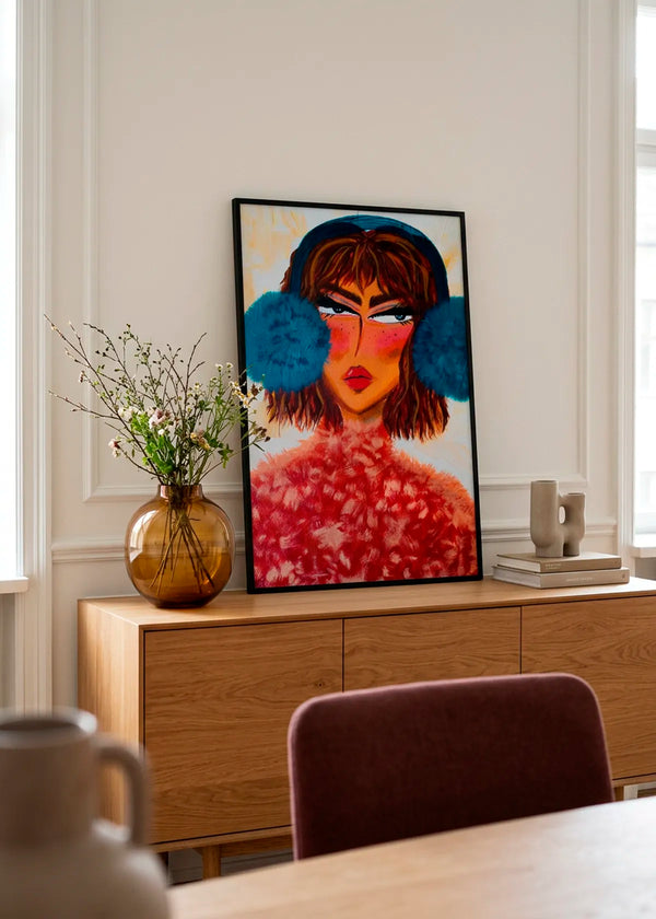 Sassy Earmuffs Portrait by Amy Straley – framed expressive portrait poster featuring a woman with bright blue earmuffs and a textured red top, styled on a warm wooden cabinet in a bright neutral interior.