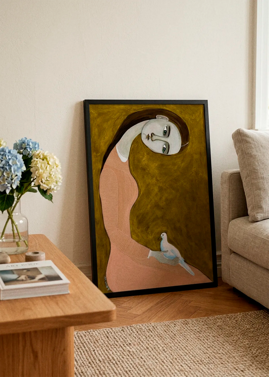 Serene Woman and Dove by Sharyn Bursic displayed in a black frame, leaning against a soft cream wall beside a beige sofa and wooden coffee table in a warm, elegant living room.