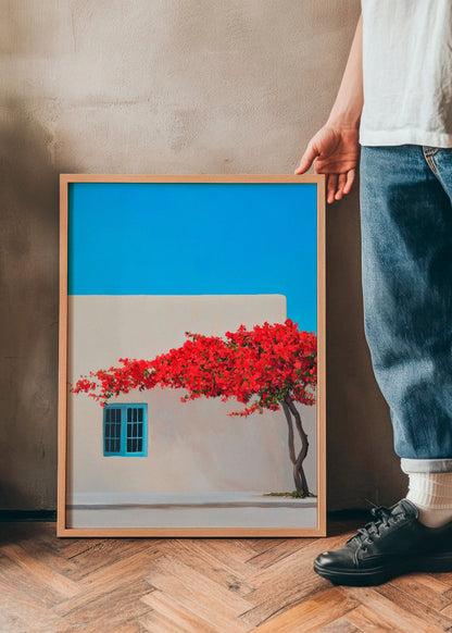 Siesta poster featuring a vibrant red bougainvillea tree against a soft beige wall and a bright blue sky. The artwork is framed in natural wood and held by a person standing on a wooden herringbone floor, creating a warm and Mediterranean-inspired atmosphere.