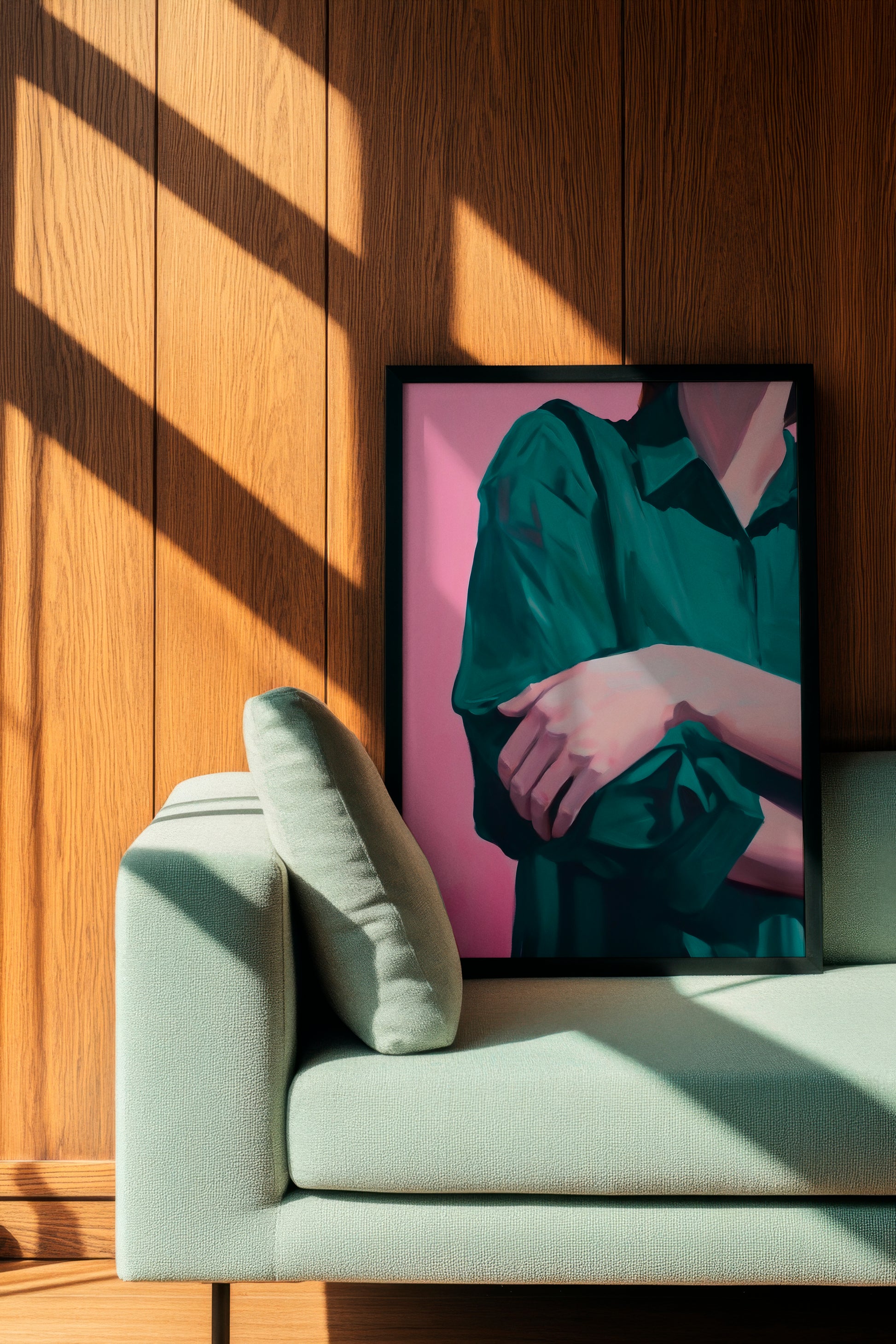 Silent Pose poster featuring a serene depiction of folded arms in a green shirt against a soft pink background, framed in black and leaning against a wooden-paneled wall beside a light green sofa. The artwork conveys calmness and introspection, perfect for minimalist and modern interiors.