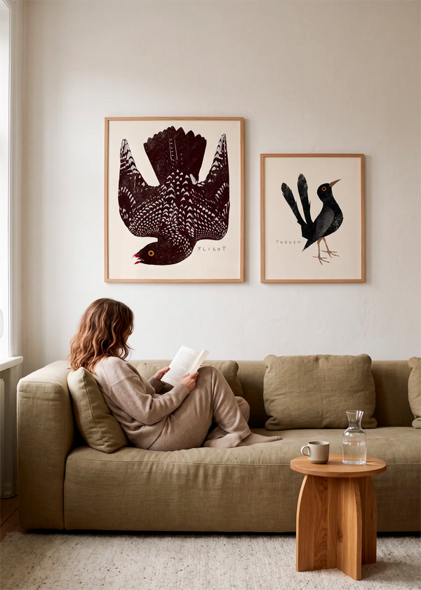 Speckled Bird Flight og Elegant Black Thrush plakater af Josephin Hermsdorf i lyse trærammer, stylet over en beige sofa i et roligt nordisk interiør.