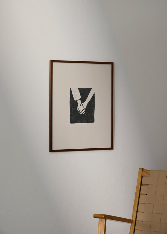 Togetherness poster from HelloPoster in dark wooden frame, minimalist black and white illustration of two hands holding, styled on a light wall with wooden chair.