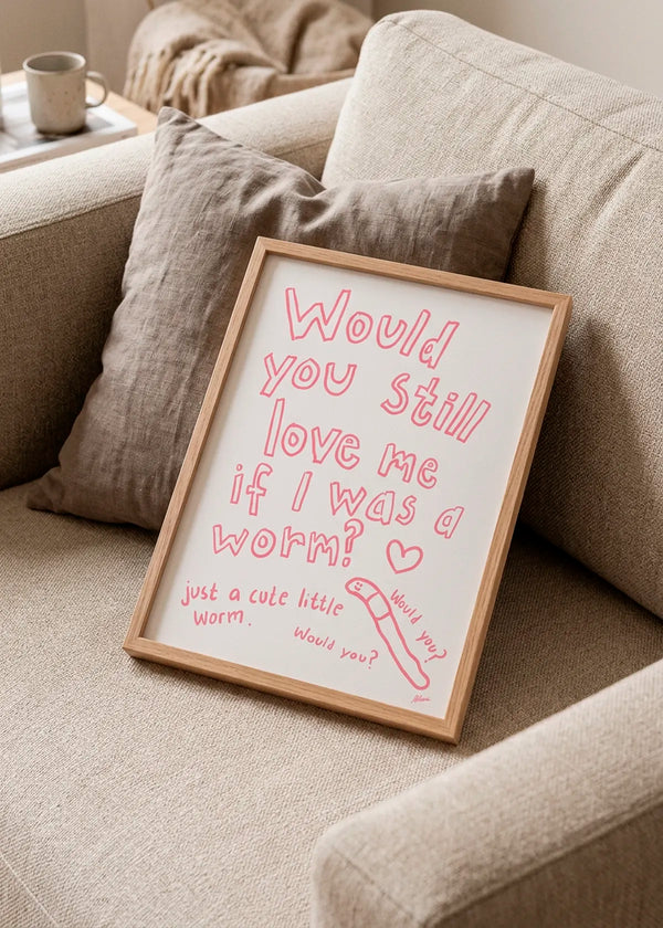 Would You? by Athene Fritsch – framed pink typographic art poster with a hand-drawn worm, styled on a neutral beige sofa with soft cushions.