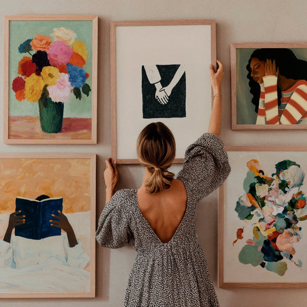 Gallery wall with Hello Poster artworks including Blossom floral print, Togetherness hand illustration, HumDrumDolly portrait, Pétale abstract flowers, and Must Read reading portrait. Styled in a warm Scandinavian interior with a woman arranging the frames.