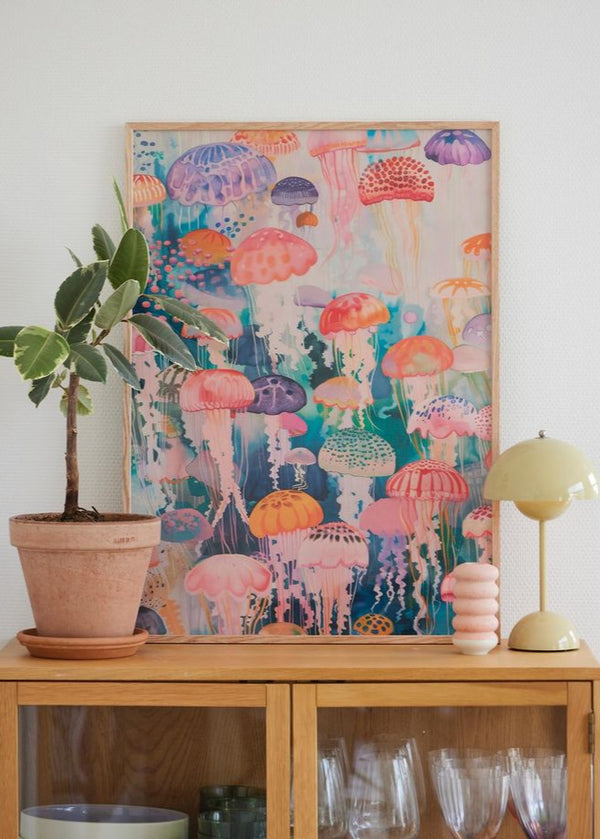 Farverig plakat med vandmænd i træramme, stylet på egetræsskab sammen med grøn lampe og stueplante.